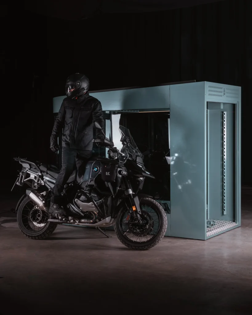 Rider standing with a motorcycle next to a MotoDome garage in a dark studio